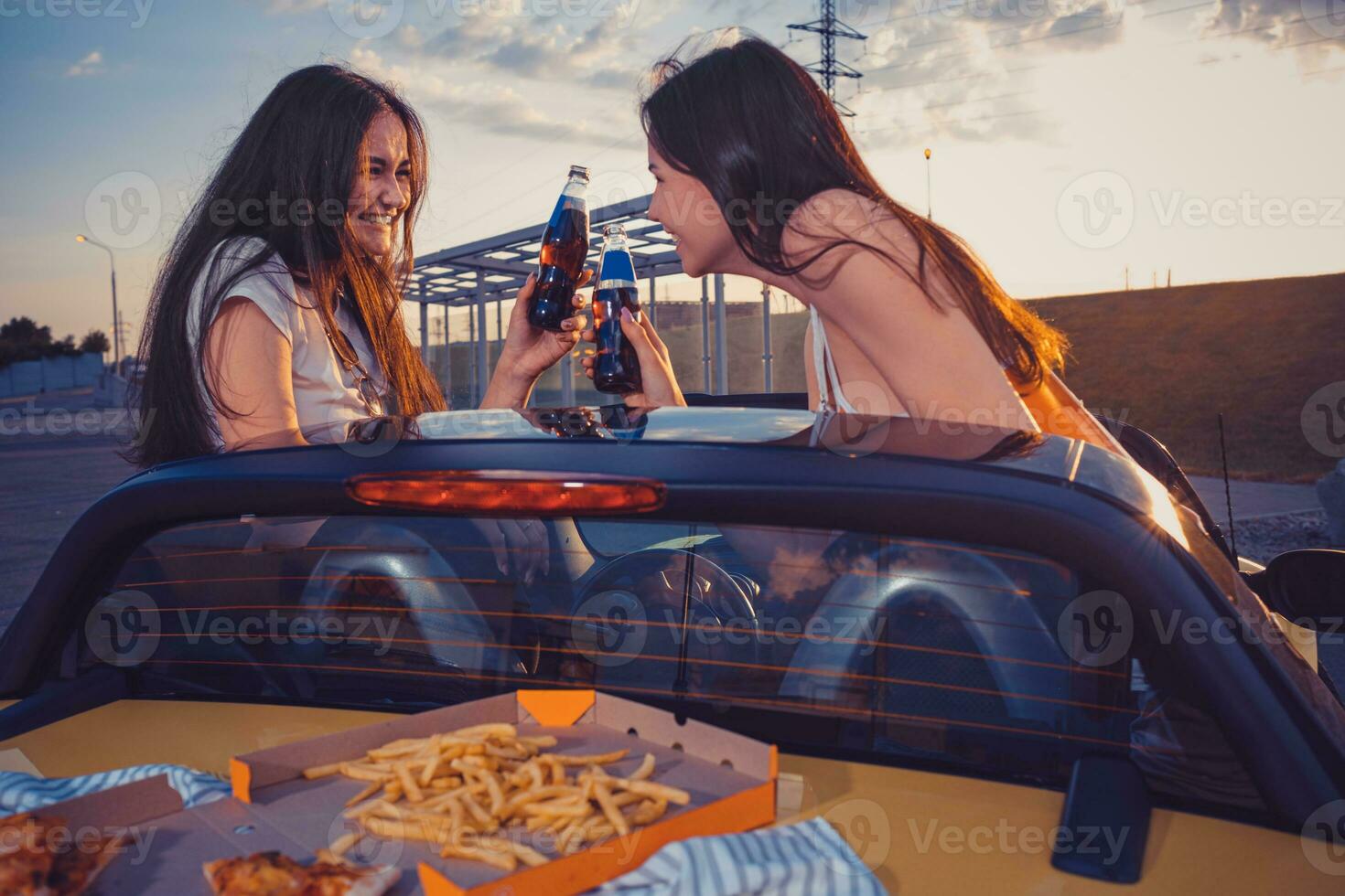 Lycklig kvinnor är leende, glädjande med soda i glas flaskor, Framställ i gul bil med franska frites och pizza på dess trunk. snabb mat. kopia Plats foto