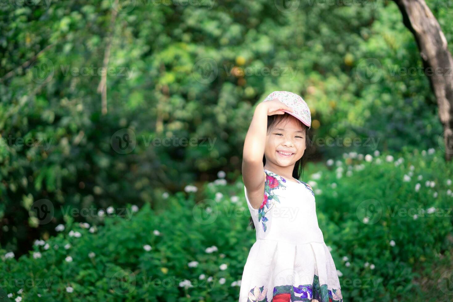 liten asiatisk flicka i klänning leende medan bär keps i de parkera foto