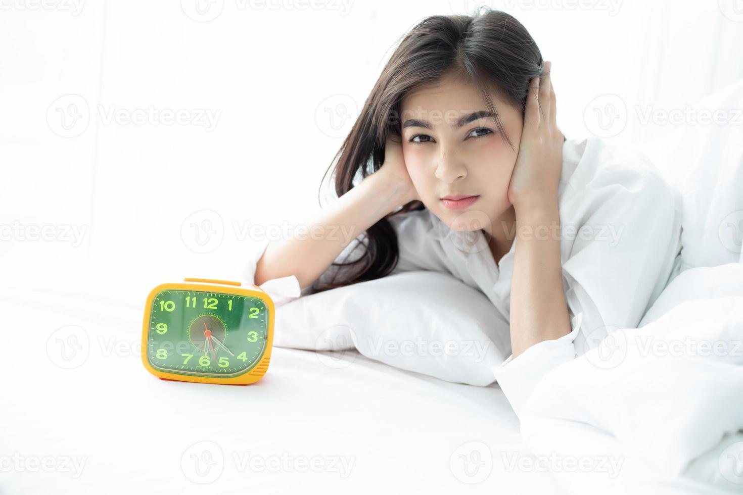 asiatisk kvinna hatar att vakna tidigt på morgonen. sömnig tjej tittar på väckarklockan och försöker gömma sig under kudden foto