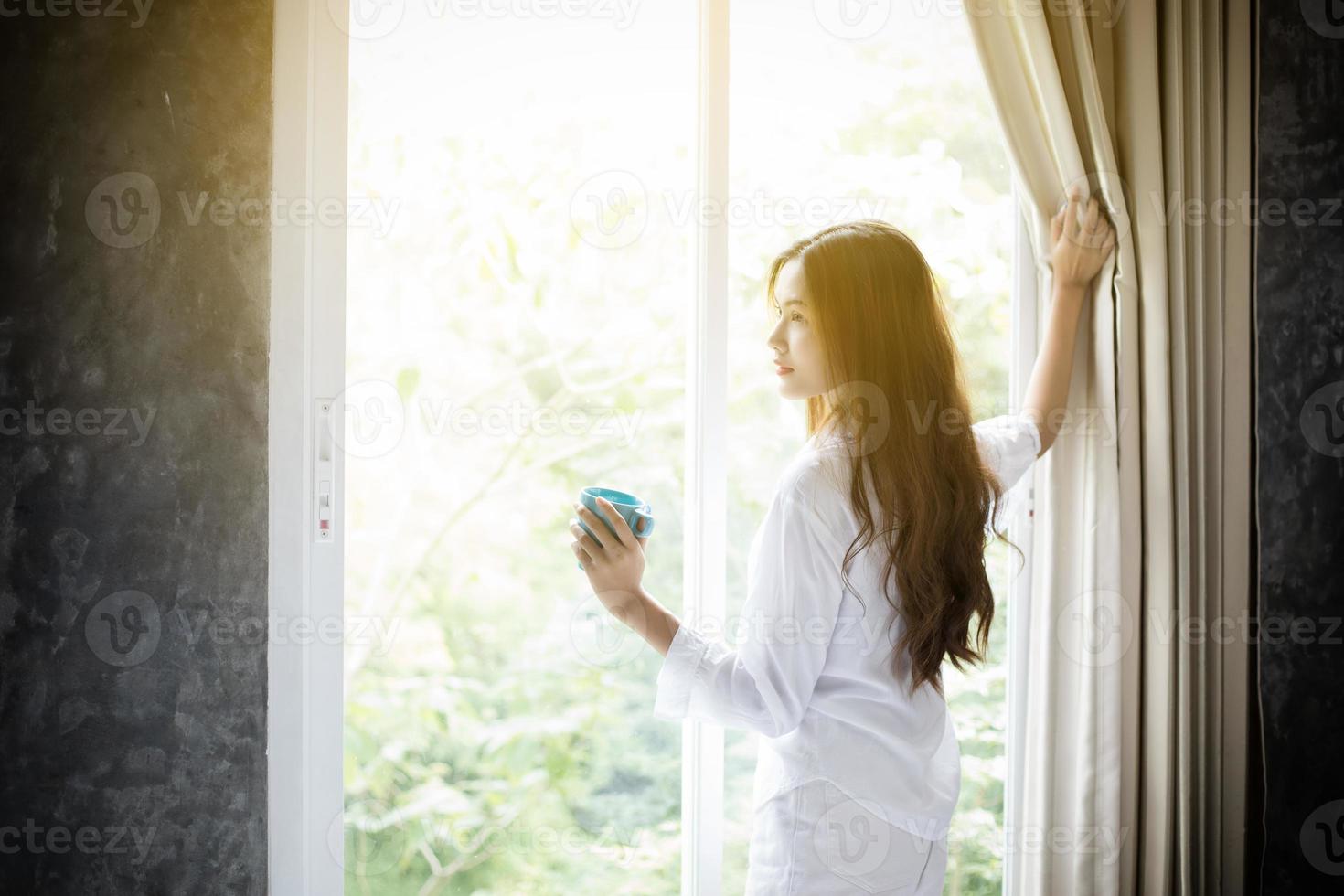 asiatiska kvinnor som dricker kaffe och vaknar i sin säng helt vilade och öppnar gardinerna på morgonen för att få frisk luft på solskenet foto