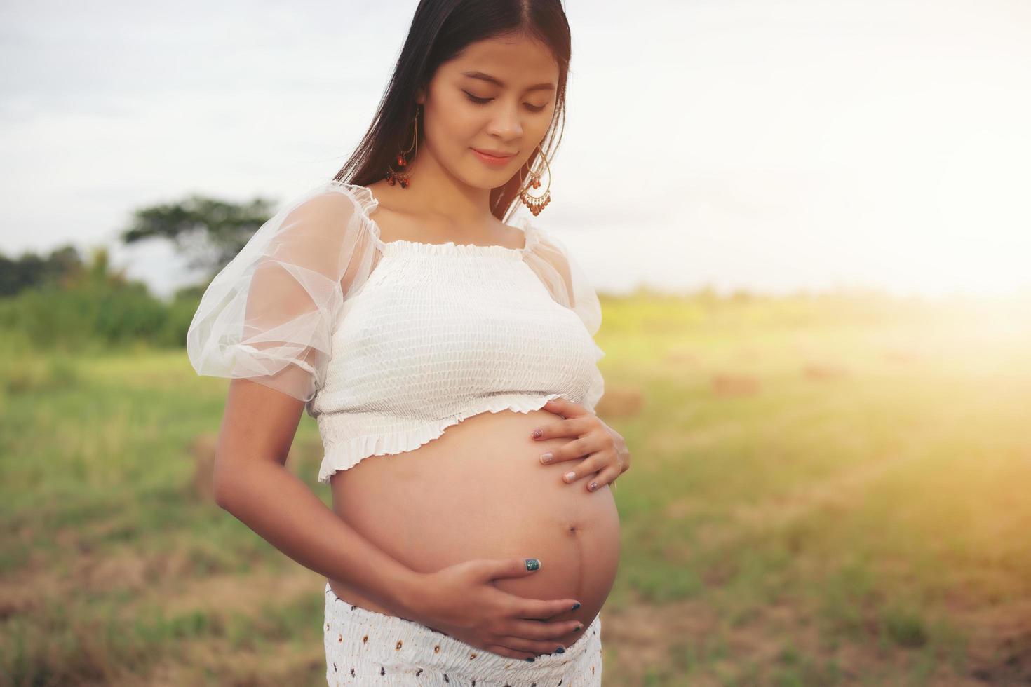 glad och stolt gravid asiatisk kvinna som tittar på hennes mage i en park vid soluppgången foto