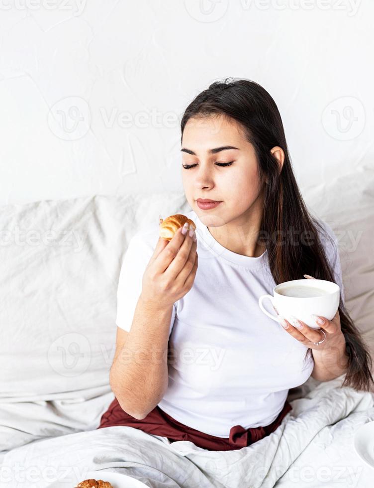 ung brunettkvinna som sitter i sängen och äter croissanter foto