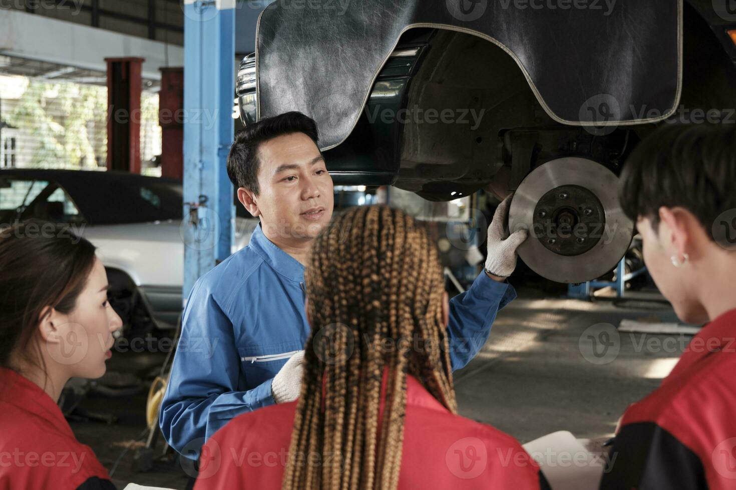 asiatisk manlig professionell bil- ingenjör handledare beskriver bil hjul och suspension reparera arbete med mekaniker arbetstagare staber team i fixera service garage, specialist yrken i bil industri. foto