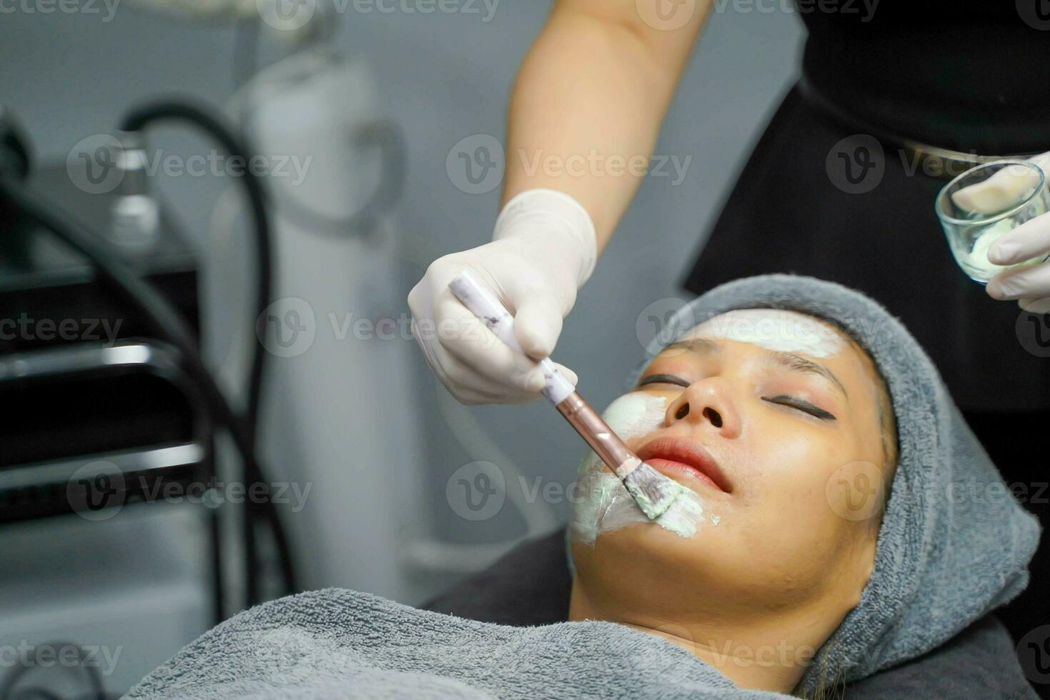 närbild kosmetolog använda sig av kosmetisk borsta applicering ansiktsbehandling vit mask grädde på kvinna klient ansikte i skönhet klinik. foto
