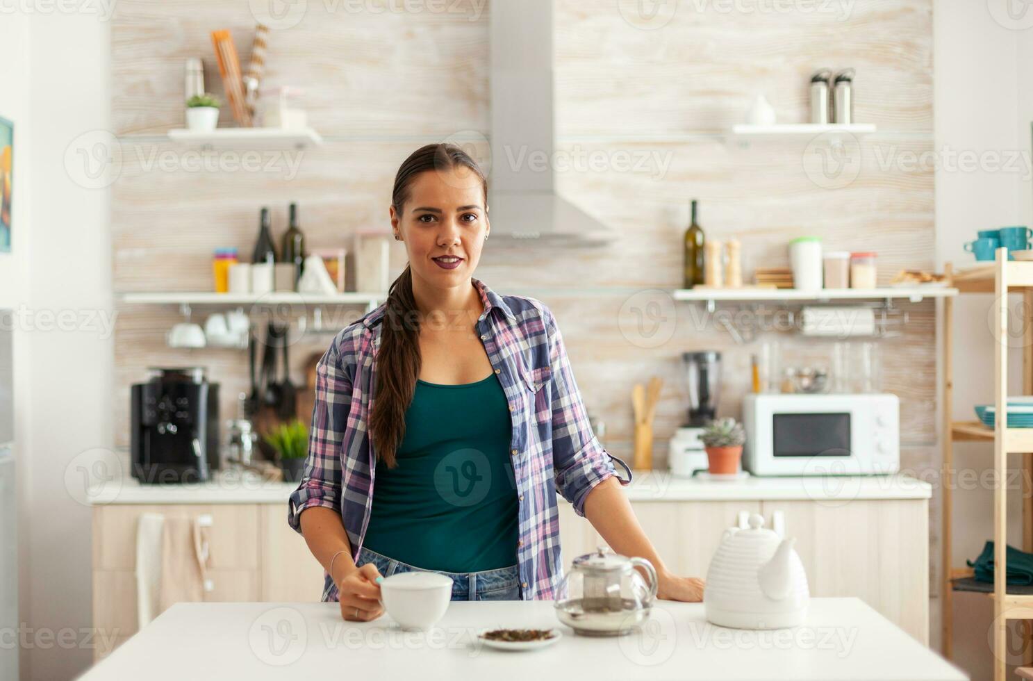 kvinna innehav kopp av te under morgon- i kök medan njuter frukost. framställning te i de morgon, i en modern kök Sammanträde nära de tabell. sätta med händer, friska ört- i pott. foto