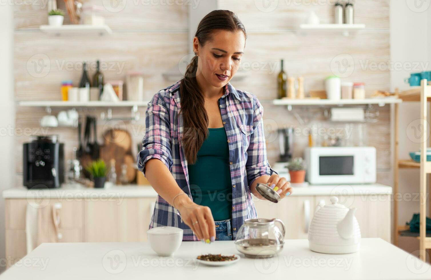 hemmafru använder sig av aromatisk örter till göra varm naturlig te för njuter frukost. framställning te i de morgon, i en modern kök Sammanträde nära de tabell. sätta med händer, friska ört- i pott. foto