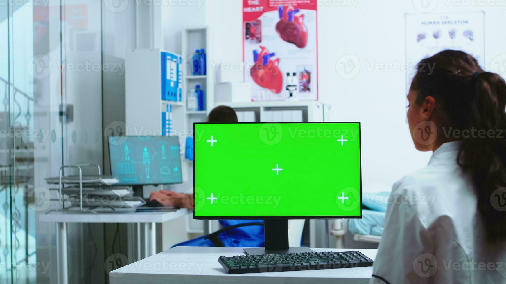 läkare använder sig av dator med grön skärm attrapp i sjukhus och assistent promenader ut av skåp bär blå enhetlig. läkare i vit täcka arbetssätt på övervaka med krom nyckel i klinik skåp till kolla upp patient diagnostisera. foto