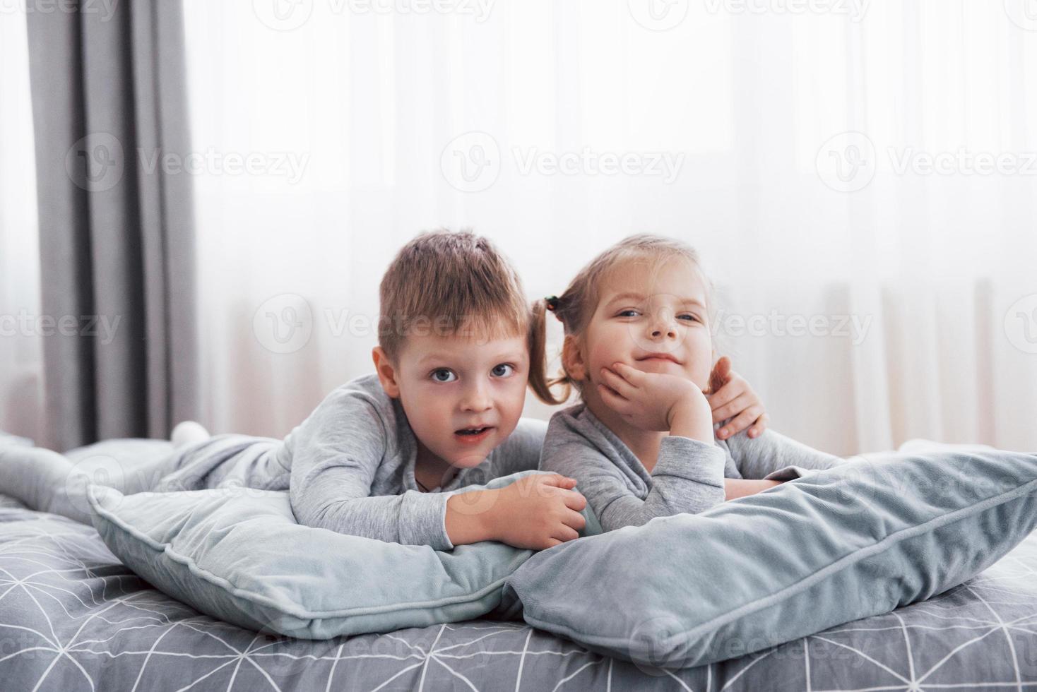 glada barn som leker i det vita sovrummet. liten pojke och flicka, bror och syster leker på sängen iklädd pyjamas. barnkammare interiör för barn. nattkläder och sängkläder för barn och småbarn. familjen hemma foto