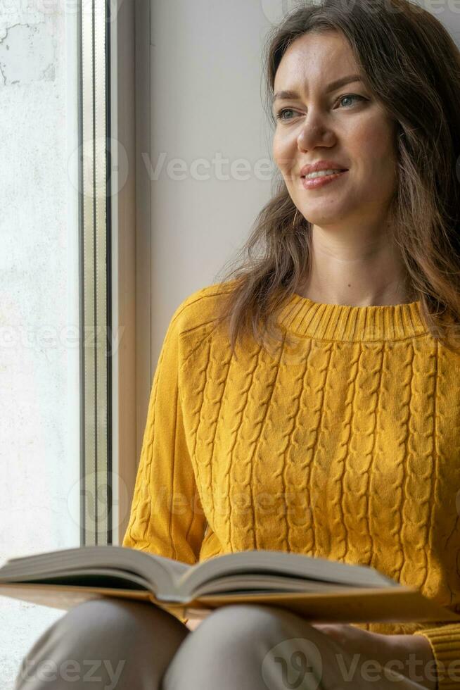 ung skön kvinna nära fönster gul stickat Tröja läsa bok foto