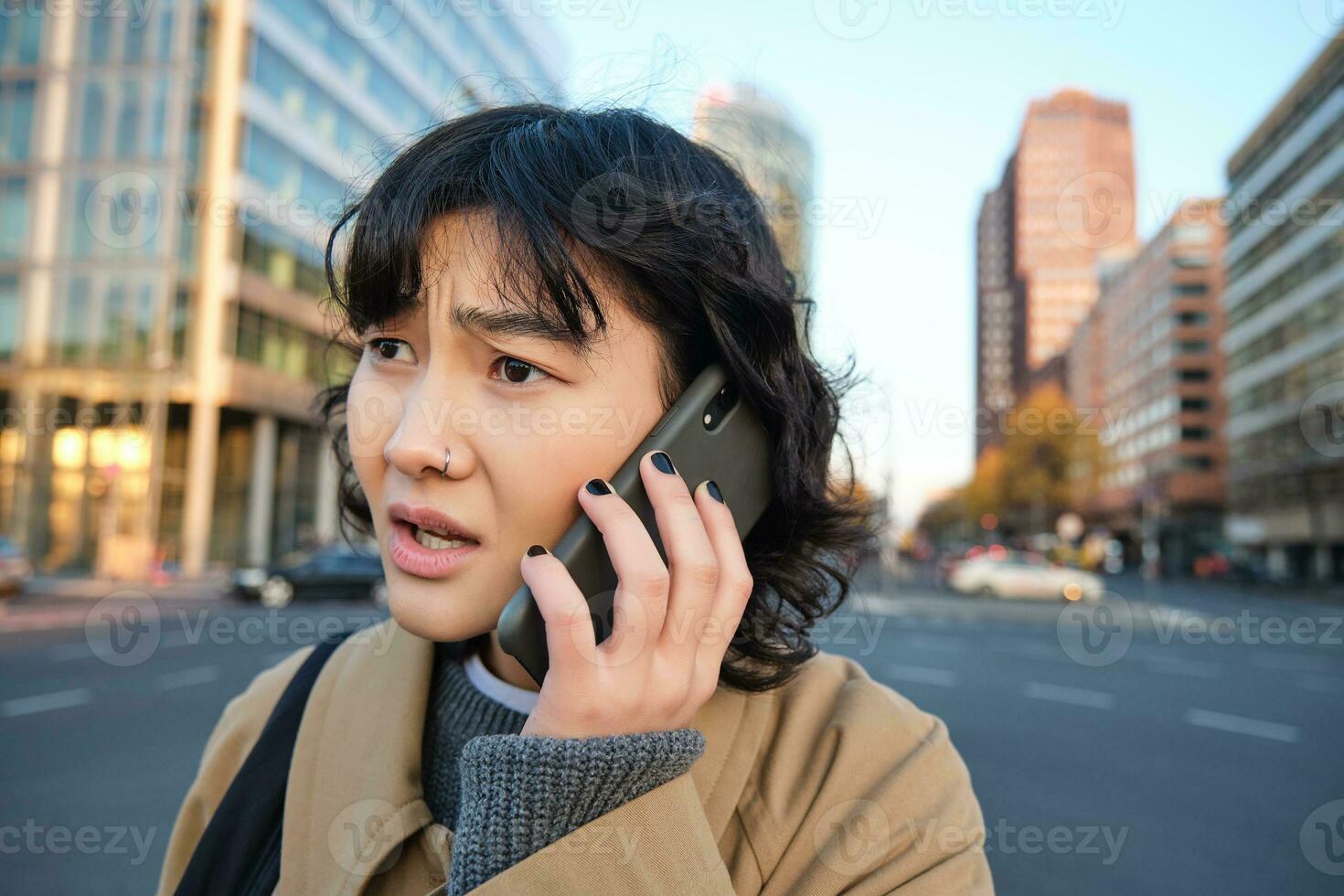 stänga upp porträtt av bekymrad asiatisk kvinna, samtal på mobil telefon och hör dålig Nyheter, utseende orolig, känner frustrerad förbi telefon konversation, står på gata foto