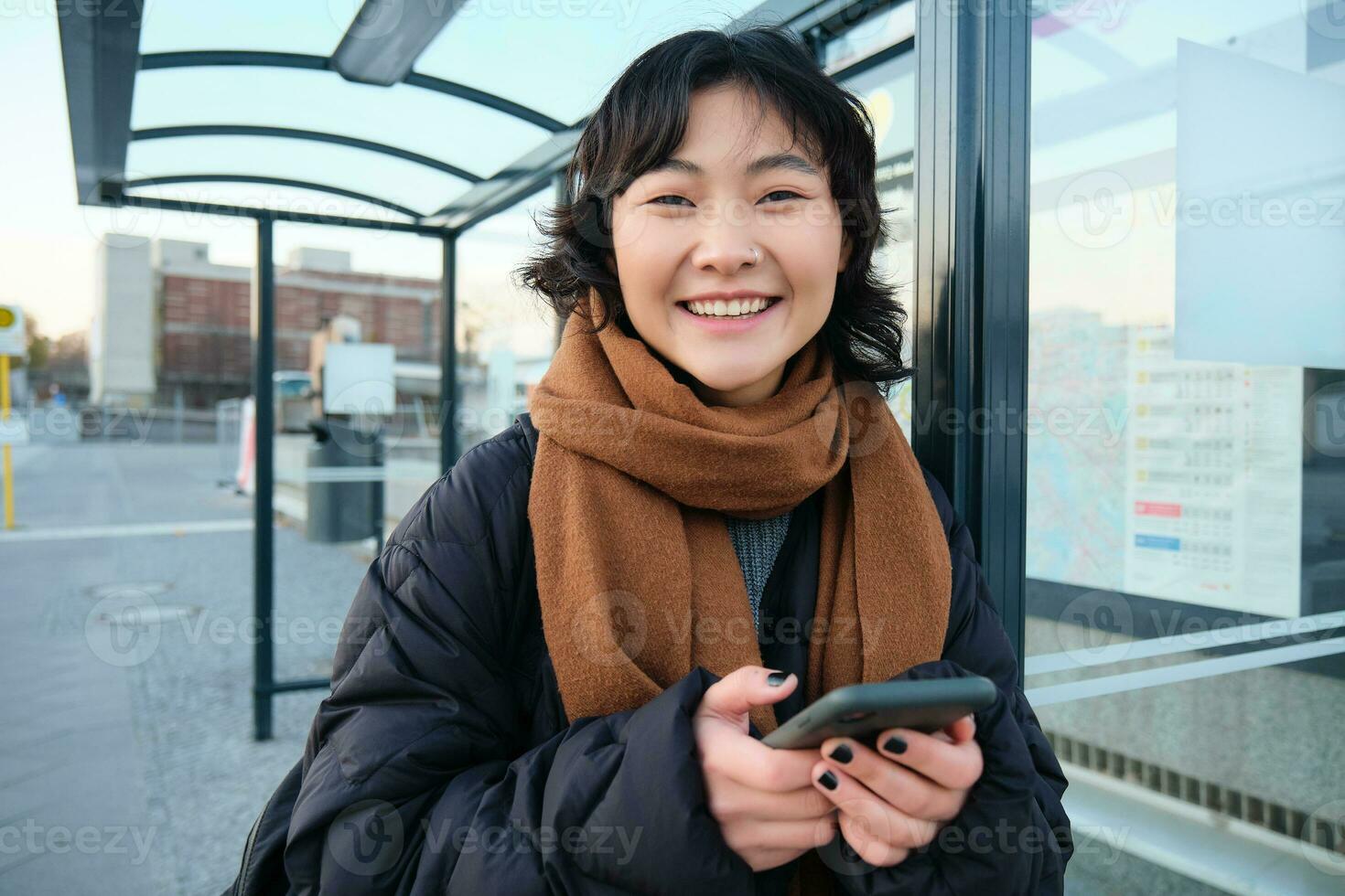 skön ung kvinna, stående på buss sluta, väntar för henne offentlig transport, använder sig av mobil telefon, använder sig av smartphone Ansökan foto