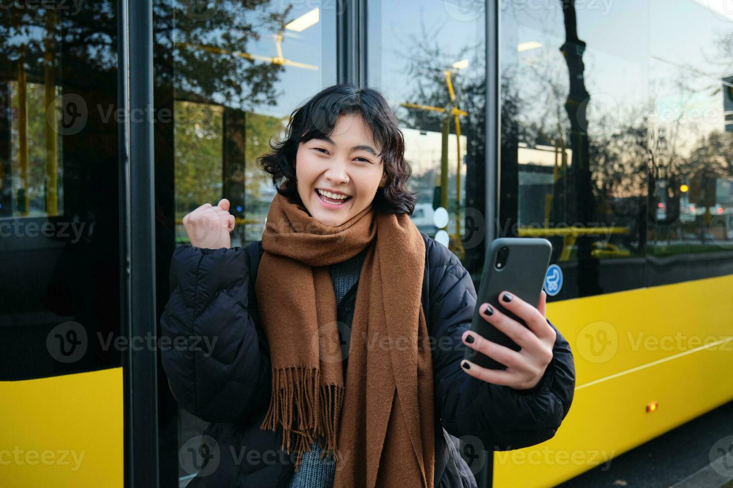 entusiastisk asiatisk kvinna, stående på buss sluta med smartphone, ser på telefon skärm med förvånad, triumferande ansikte, vinnande, höra bra Nyheter på video chatt foto