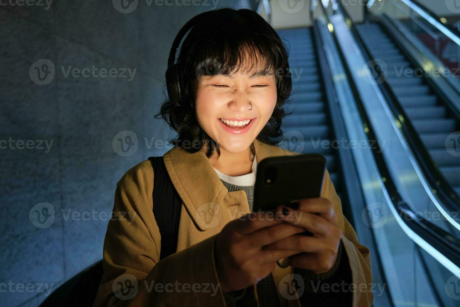 skrattande söt koreanska flicka på rulltrappa, gående ner till rör, underjordisk eller metro, använder sig av mobil telefon, tittar på smth rolig på smartphone app foto