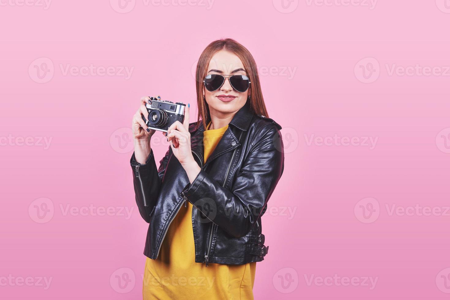 mode look, ganska cool ung kvinna modell med retro film kamera bär en svart jacka, mot rosa bakgrund med kopia utrymme foto