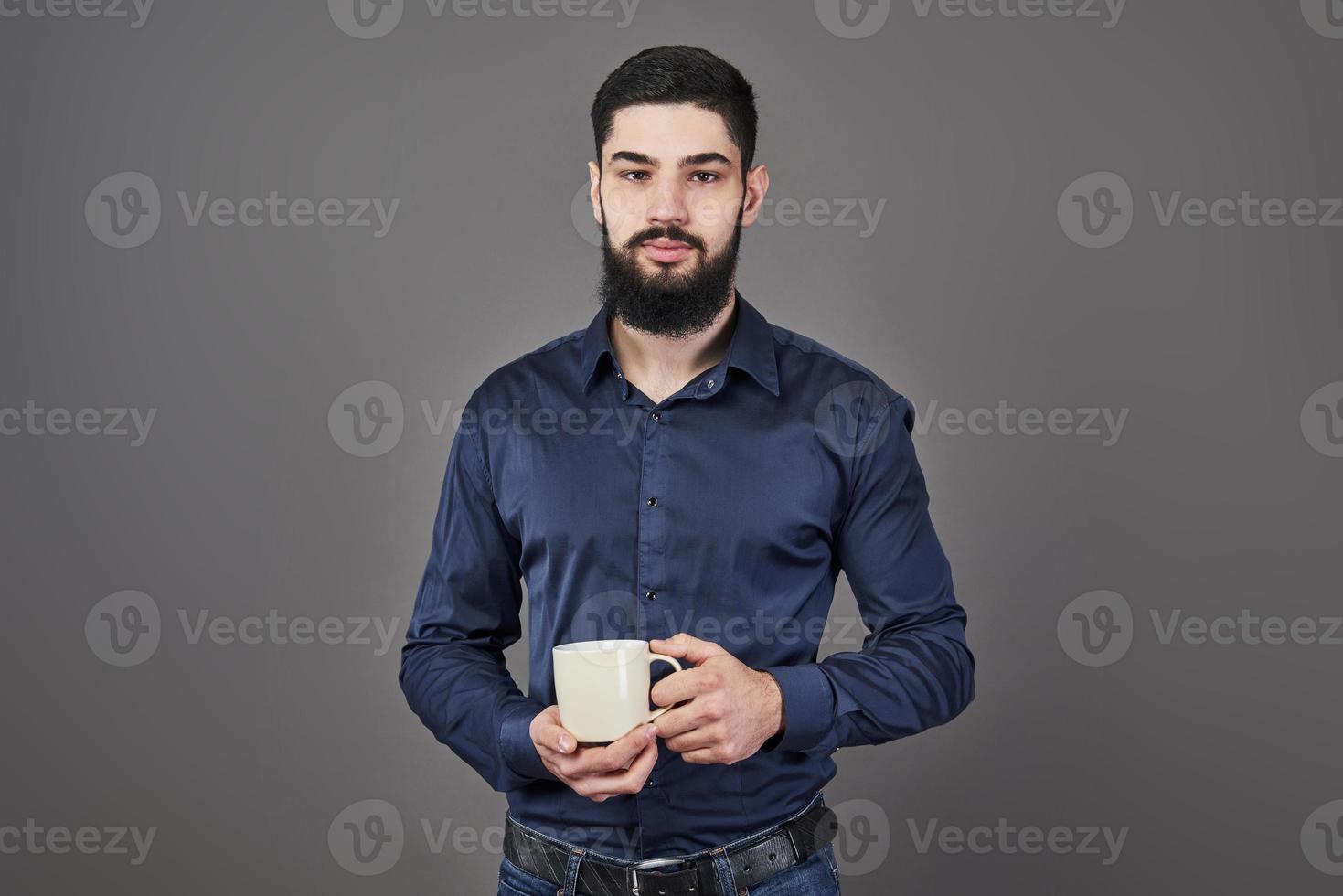 stilig skäggig man med snyggt hårskägg och mustasch på allvarligt ansikte i skjorta som håller vit kopp eller mugg som dricker te eller kaffe i studion på grå bakgrund foto