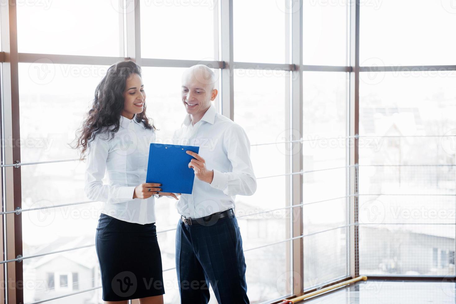 affärsman och affärskvinna som studerar ett diagram på tallriken och pappersdokument vid fönstret på bakgrunden av stadskontoret på en hög våning foto