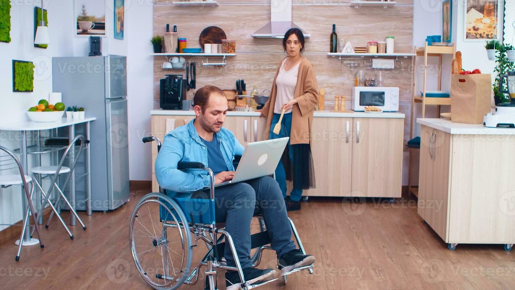 handikappade entreprenör i rullstol arbetssätt på bärbar dator medan fru är framställning måltid i kök. man med förlamning handikapp handikapp handikappade svårigheter få hjälp för rörlighet från kärlek och relation foto