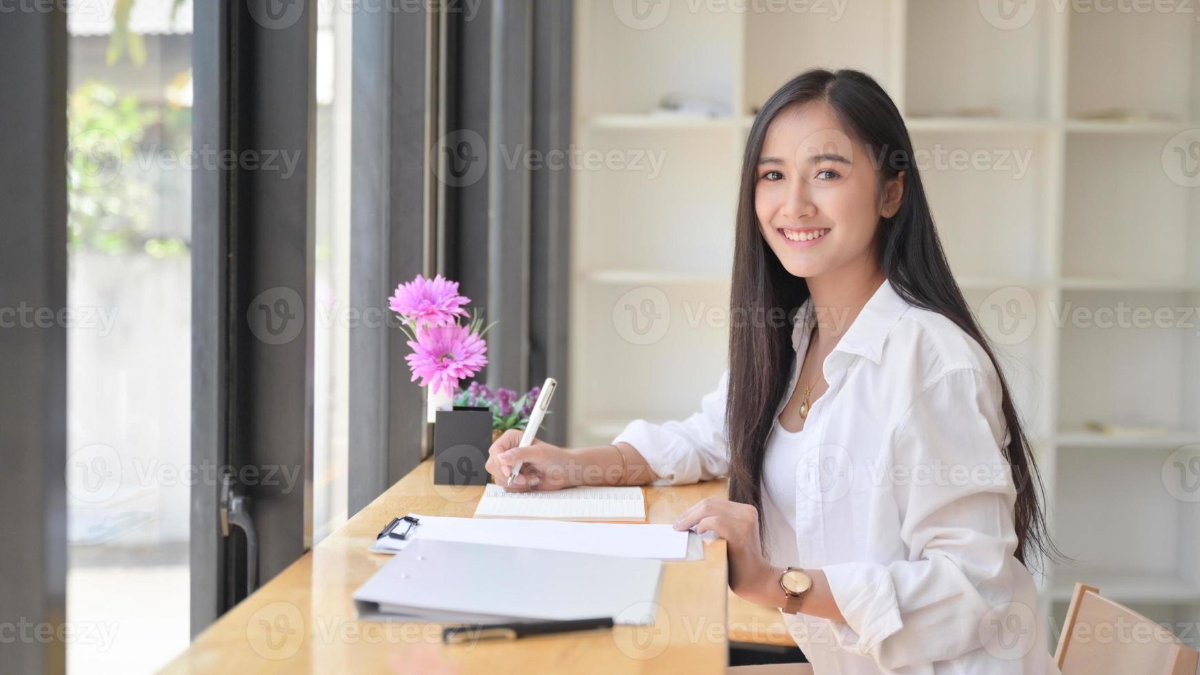 asiatisk kvinna som bär en vit klänning som håller en penna för att skriva anteckningar på ett bekvämt kontor. hon tittade på kameran och log. foto