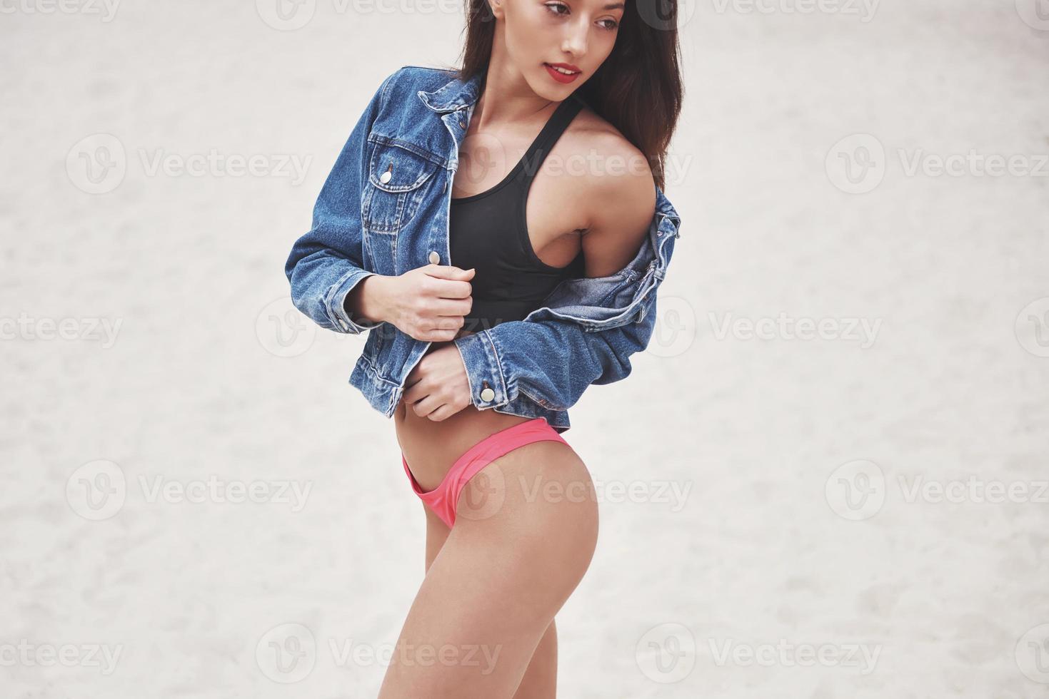 vacker smal lyxflicka i bikini och jeansjacka på sandstranden på en tropisk ö. sexig solbränd kropp och perfekt figur foto