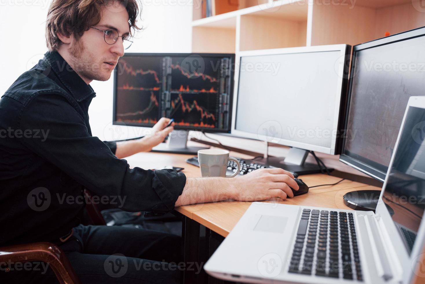 stressig dag på kontoret. ung affärsman håller händerna på ansiktet medan han sitter vid skrivbordet på kreativa kontor. börshandel forex finans grafiskt koncept foto