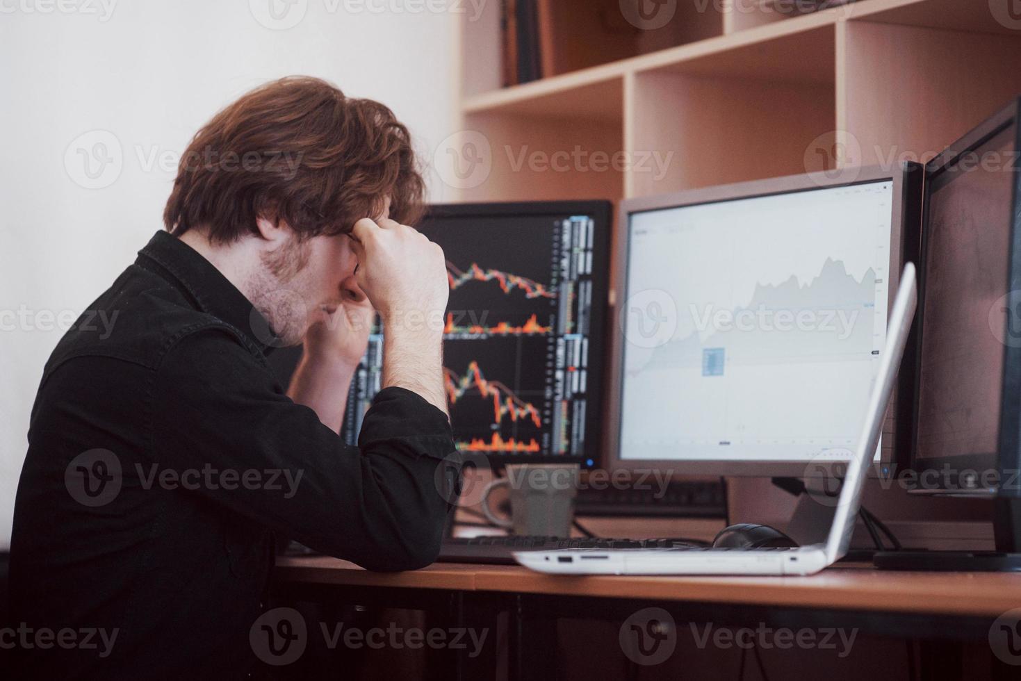 stressig dag på kontoret. ung affärsman håller händerna på ansiktet medan han sitter vid skrivbordet på kreativa kontor. börshandel forex finans grafiskt koncept foto