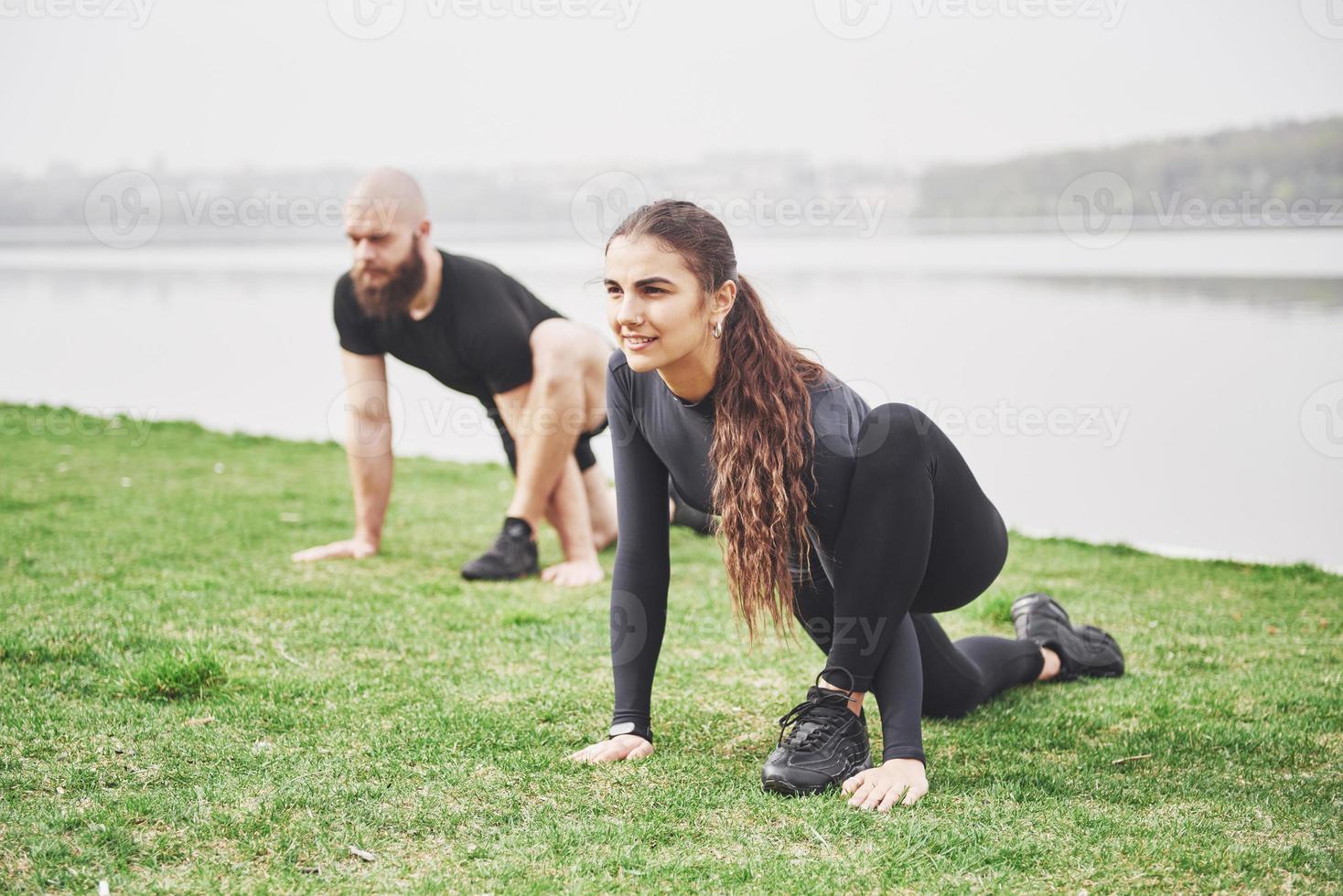 fitness par som sträcker sig utomhus i parken nära vattnet. ung skäggig man och kvinna som tränar tillsammans på morgonen foto