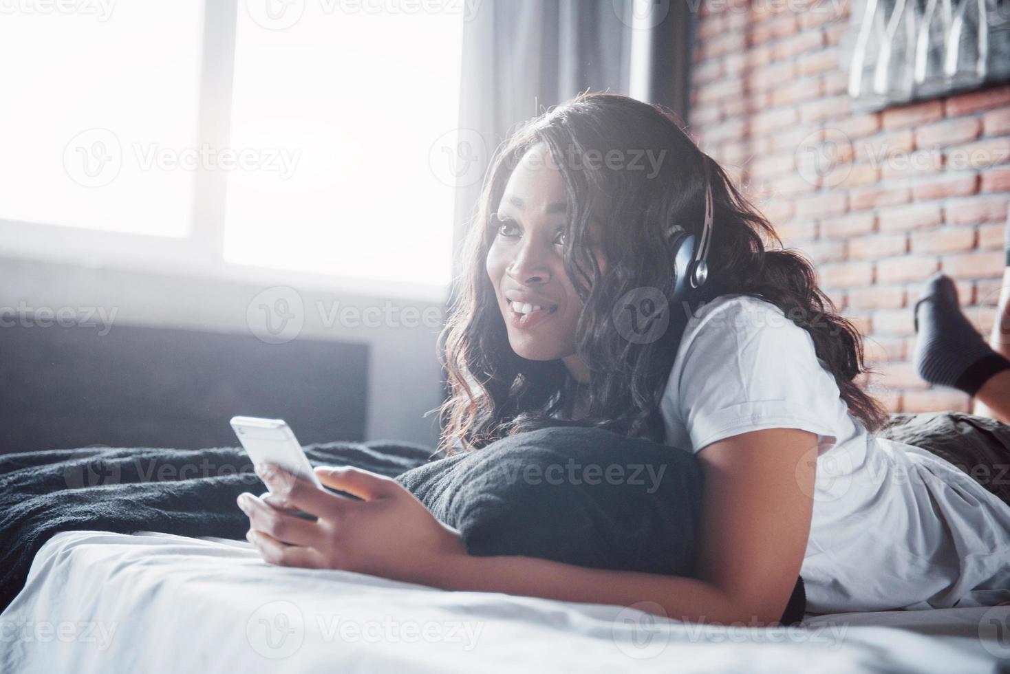 vacker afrikansk flicka i sängkläder leende lyssna på musik i hörlurar som ligger på sängen foto