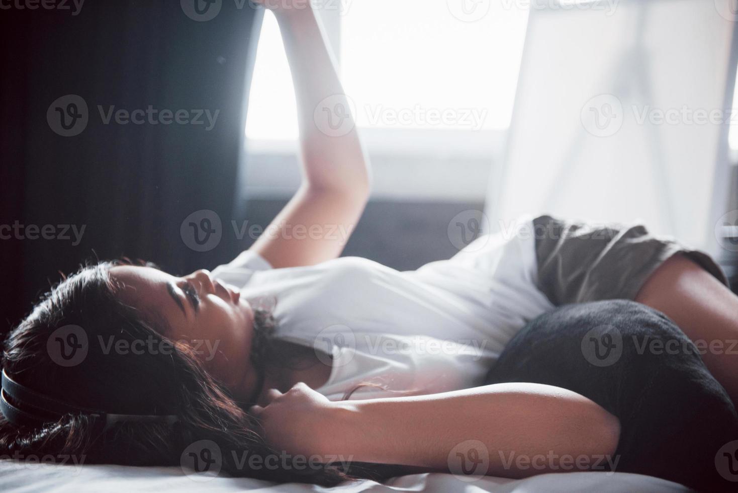 porträtt av vacker kvinna som vaknar i sin säng och tittar in i telefonen. kolla sociala nätverk, skicka sms. flickan bär en t-shirt foto