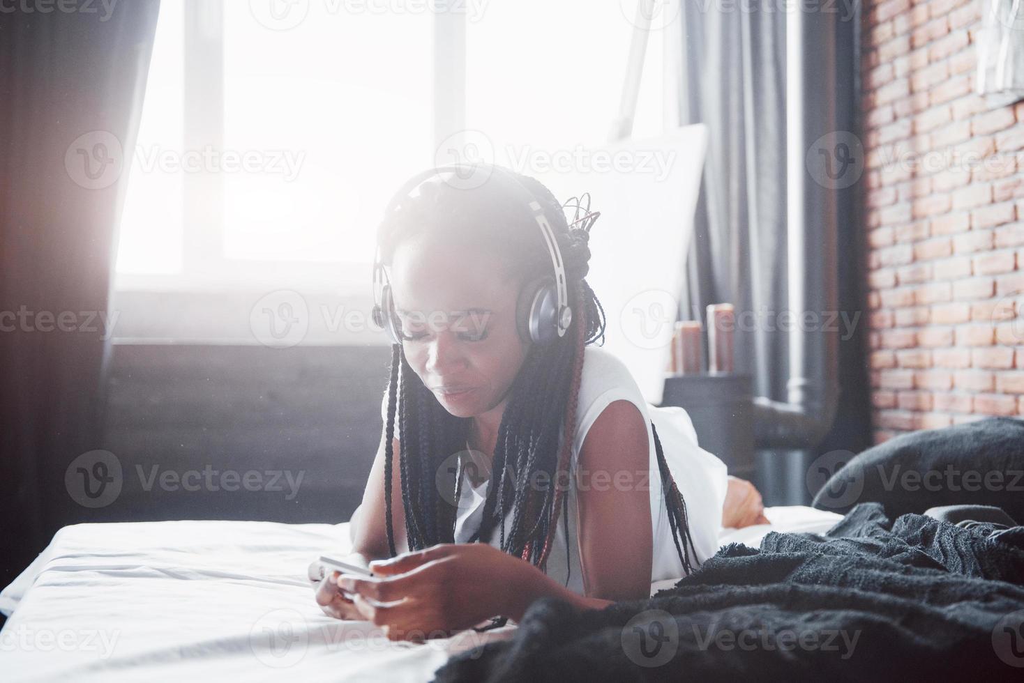 vacker afrikansk flicka i sängkläder leende lyssna på musik i hörlurar som ligger på sängen foto