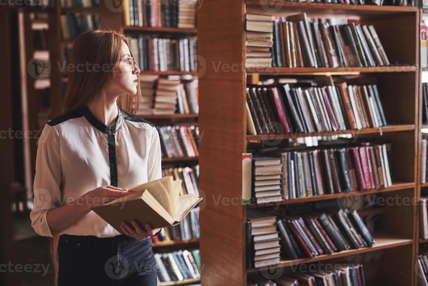 ung attraktiv studentbibliotekarie som läser en bok mellan bibliotekets bokhyllor foto