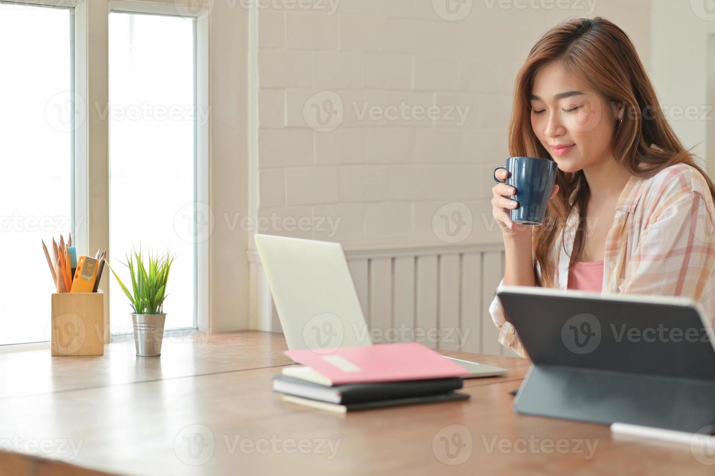 asiatisk studentflicka som håller en kaffekopp och använder en bärbar dator arbetar med ett projekt för att avsluta sina studier. foto