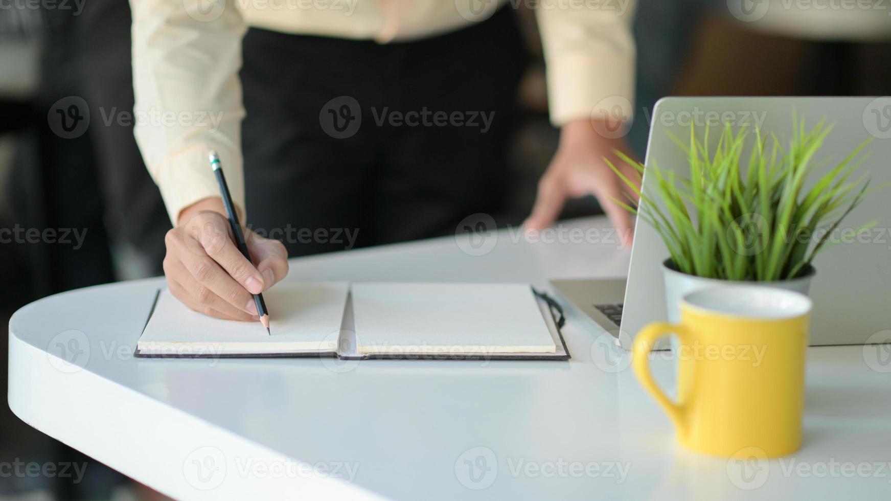 handen skriver en anteckning på en anteckningsbok med en kaffemugg och en bärbar dator på skrivbordet. foto