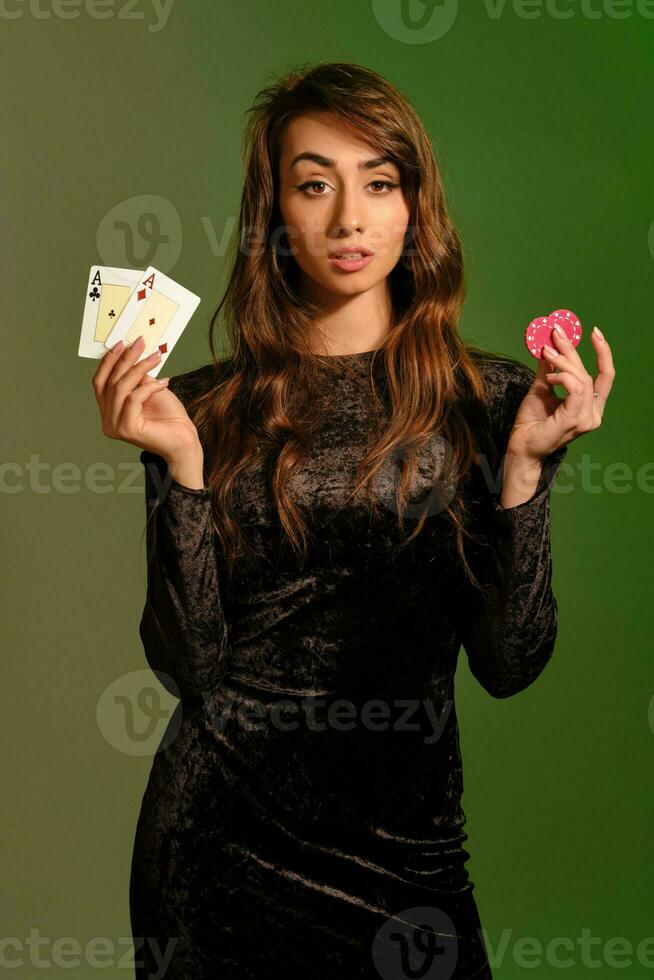 brunett flicka i svart klänning som visar två röd pommes frites och ess, Framställ mot färgrik studio bakgrund. spelande, poker, kasino. närbild. foto