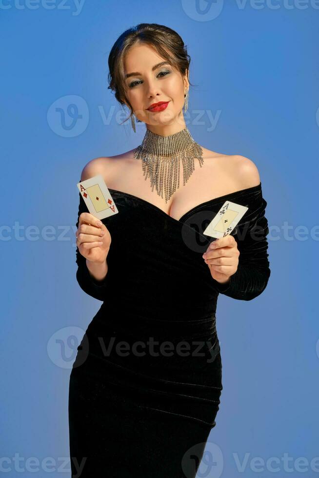 brunett kvinna med bar axlar, i svart klänning och Smycken. leende, som visar två spelar kort Framställ på blå bakgrund. poker, kasino. närbild foto