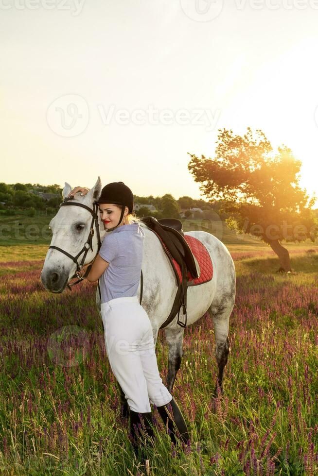 jockey ung flicka petting och kramas vit häst i kväll solnedgång. Sol blossa foto