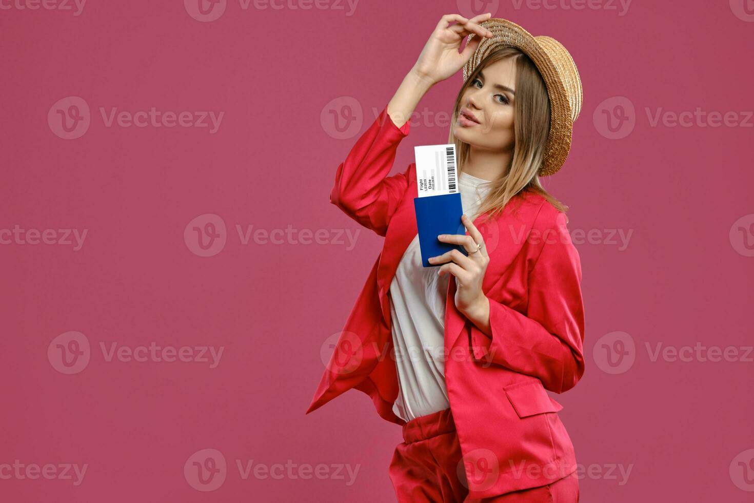 blond lady i sugrör hatt, vit blus och röd byxdräkt. leende, innehav pass och biljett medan Framställ mot rosa studio bakgrund. närbild foto