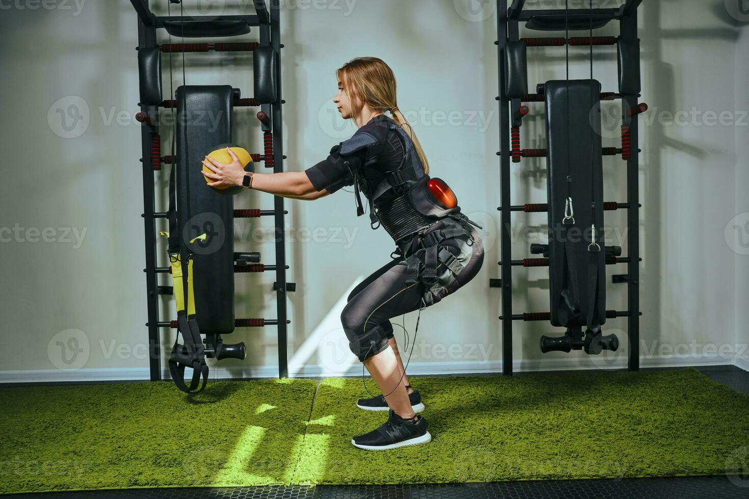 skön blond kvinna bär i sensorer och bälten, fäster med kardborreband är praktiserande ems kondition i en Gym, byggnad upp muskler med en viktad boll. modern snäll av sport. foto