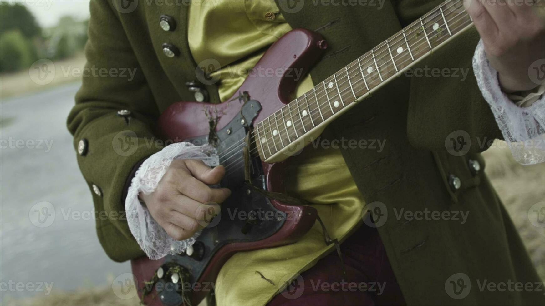 stänga upp för man med en lila elektrisk gitarr spelar i gammal fashioned kostym av rokoko- sätt på vissnade gräs bakgrund. stock. konstnär i gammal kläder spelar gitarr med hängande kelp. foto