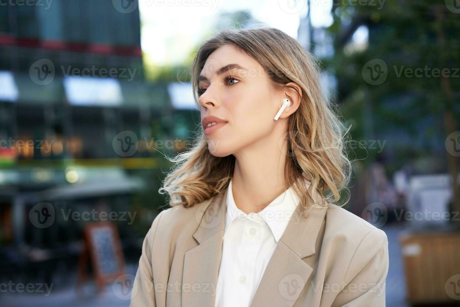 stänga upp porträtt av blond affärskvinna, självsäker företags- kvinna i hörlurar och beige kostym, Framställ utomhus på gata av stad Centrum foto