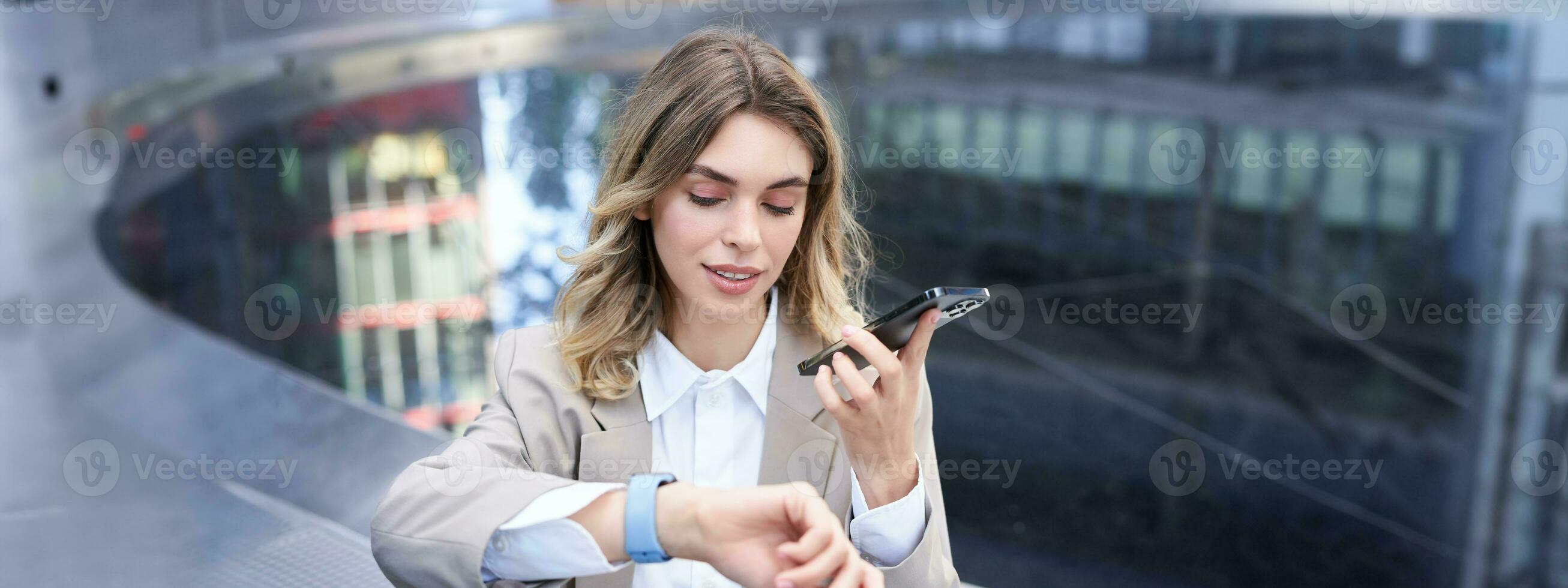 affärskvinna checkar tid på digital Kolla på och uppgifter röst meddelande, ordnar en möte, sitter utanför i stad Centrum foto