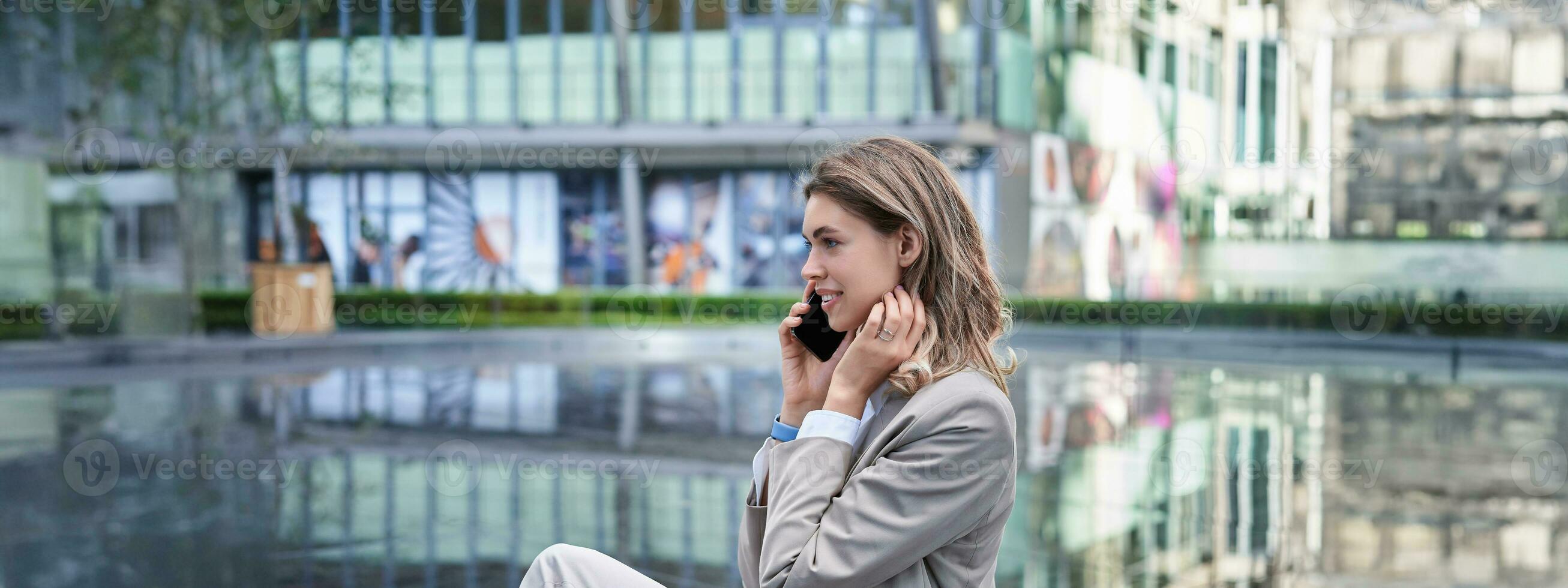 ung framgångsrik affärskvinna Sammanträde utanför kontor byggnad och talande på mobil telefon, bär kostym och hög hälar foto