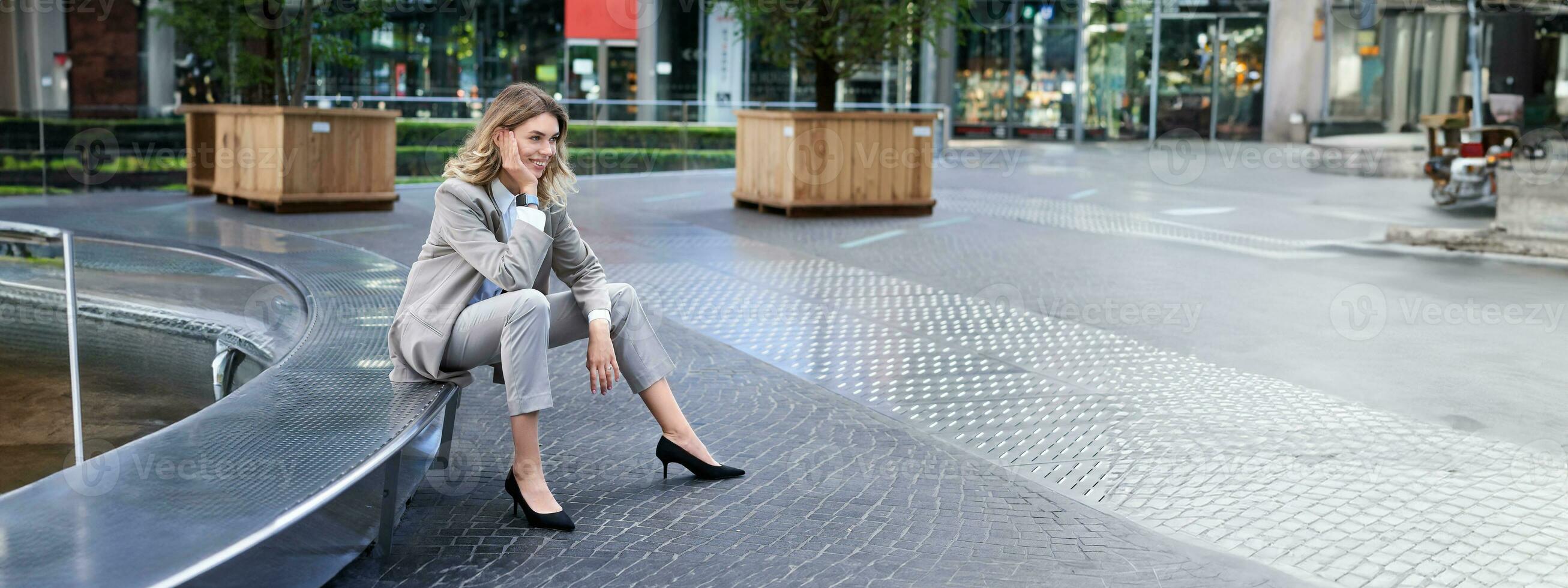 leende kvinna entreprenör i kostym, lady på henne lunch ha sönder väntar för någon nära fontän på tömma gata foto