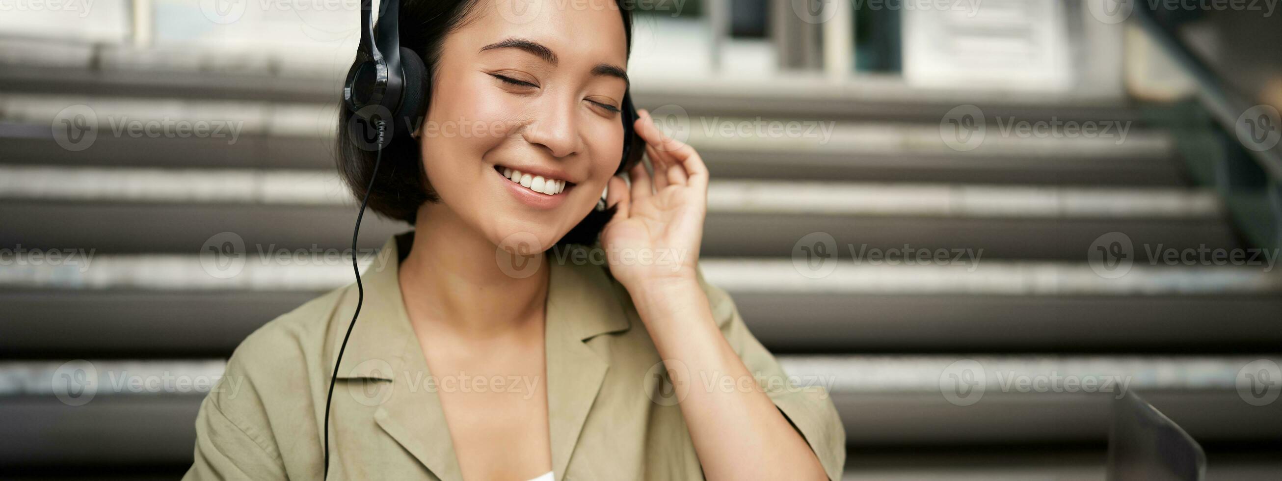 Lycklig asiatisk flicka lyssnar till musik i hörlurar, ler, sitter utomhus på trappa foto