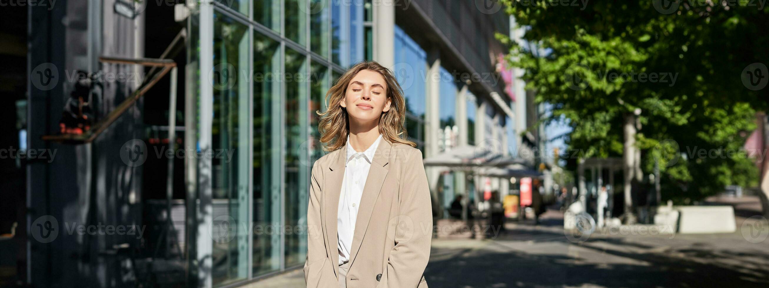 porträtt av ung skön företags- kvinna i beige kostym, leende och ser självsäker, stående på gata utanför kontor foto