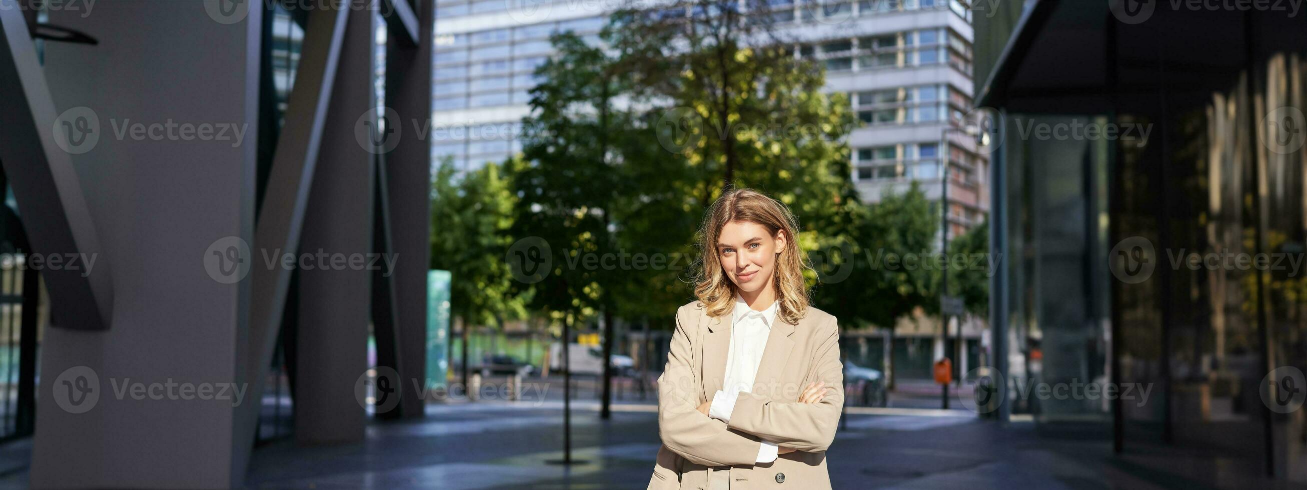 porträtt av leende affärskvinna i företags- Kläder, ser självsäker och Lycklig, bär beige kostym, stående utomhus på gata, utanför kontor foto