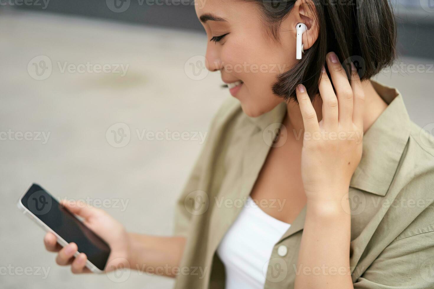 stänga upp porträtt av asiatisk flicka, ser på mobil skärm, lyssnande musik i hörlurar. kvinna med hörlurar promenader på gata foto