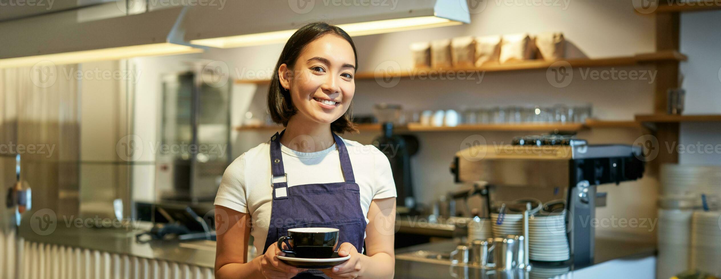 porträtt av leende asiatisk kvinna i förkläde, Barista ger du kopp av kaffe, arbetssätt i Kafé, tjänande drycker foto