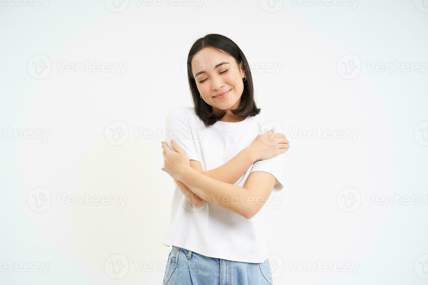 själv kärlek. Lycklig leende asiatisk kvinna, kramar själv, står över vit bakgrund foto