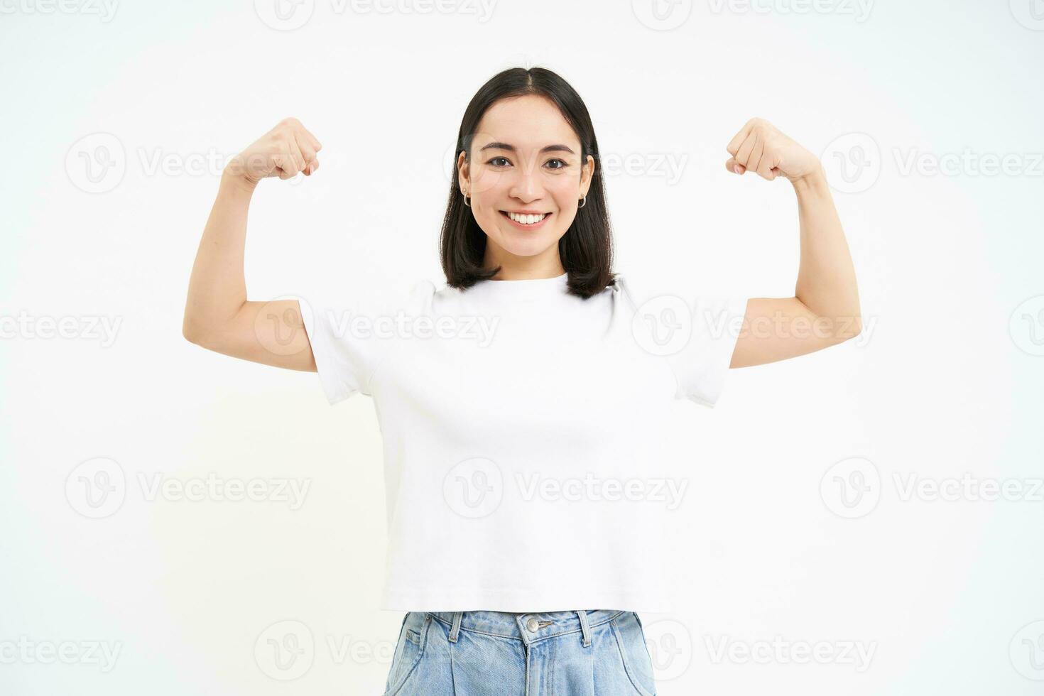 porträtt av ung asiatisk kvinna, böjning muskler, som visar biceps och leende med förtroende, ser stark, stående över vit bakgrund foto