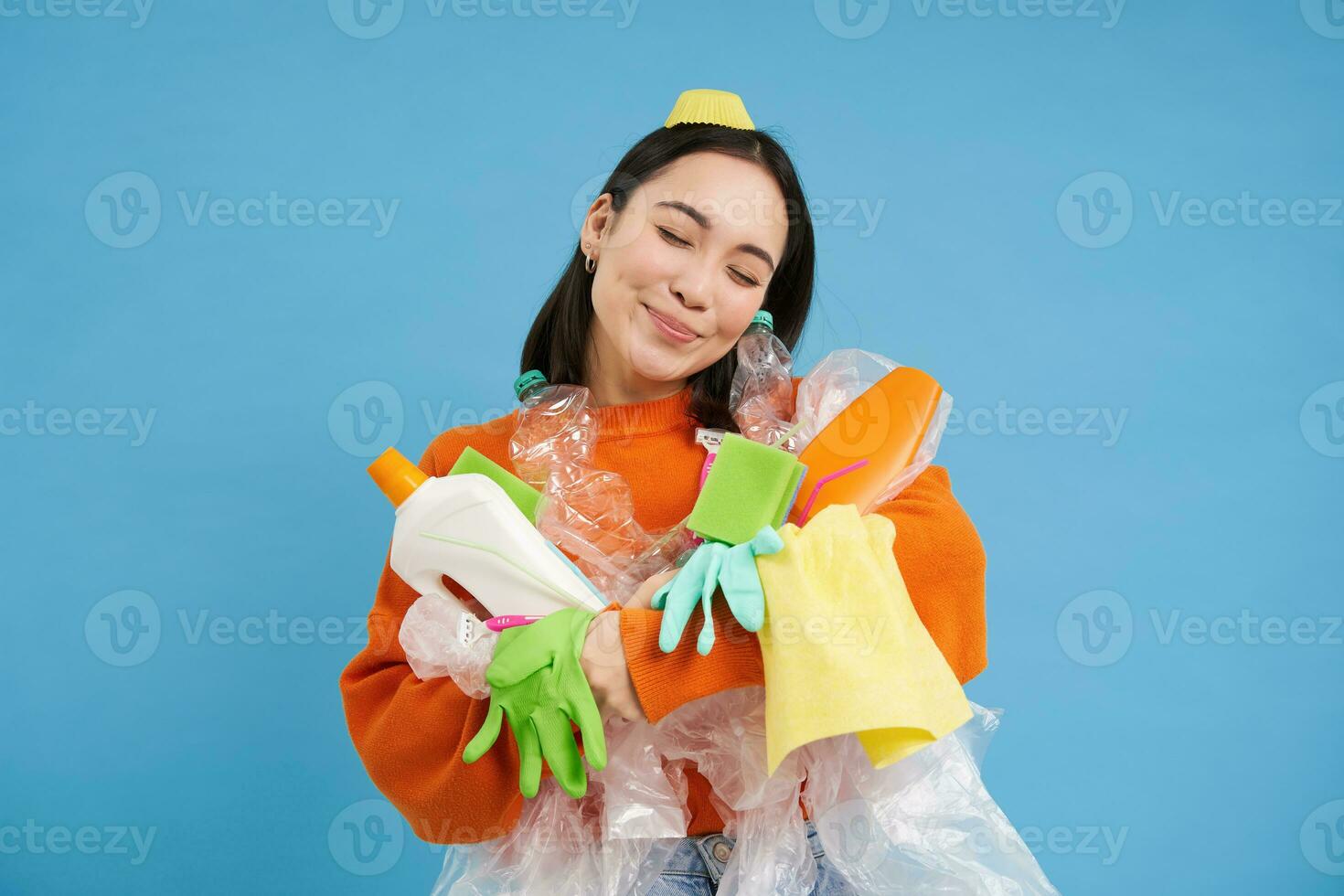 porträtt av leende asiatisk kvinna kramas tömma plast flaskor och återvinningsbar sopor, ekoaktivist gillar till återvinna, blå bakgrund foto